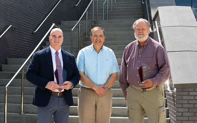 Three men stand at the base of an outdoor staircase. Two men hold trophies.